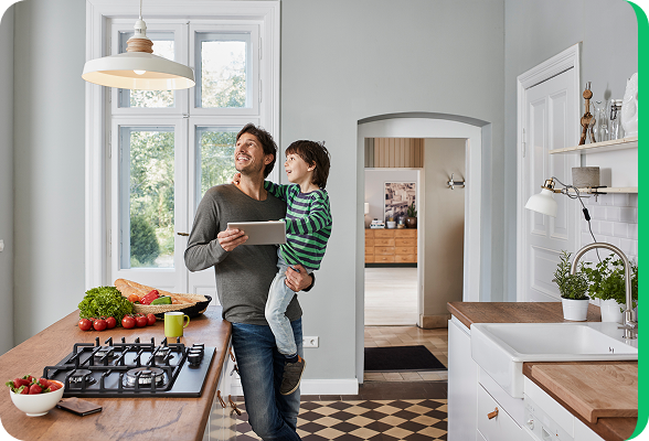 Father holding a child in a bright kitchen while looking at a tablet, illustrating managing energy consumption at home.